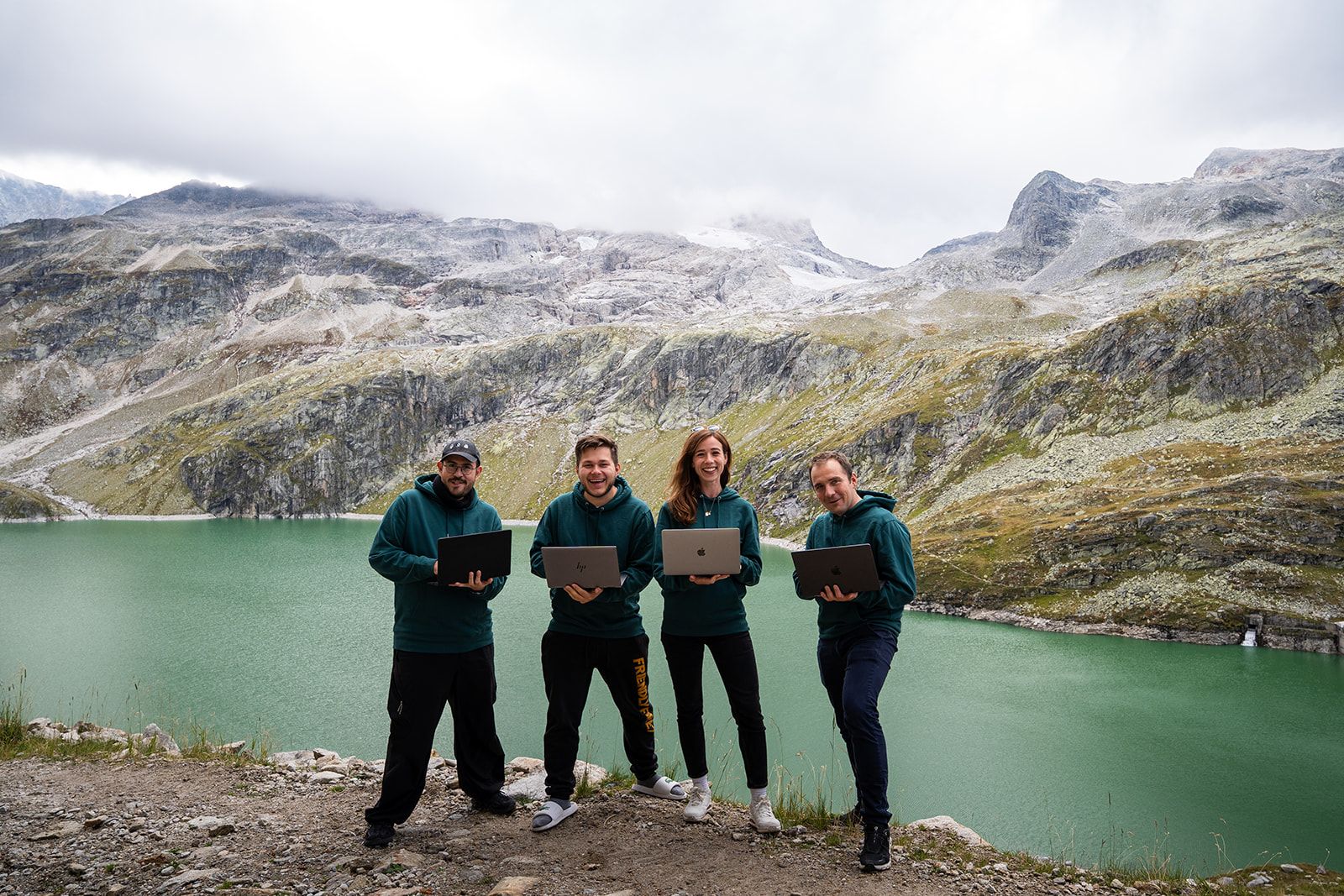 The four team members in front of a mountain lake, each holding a laptop